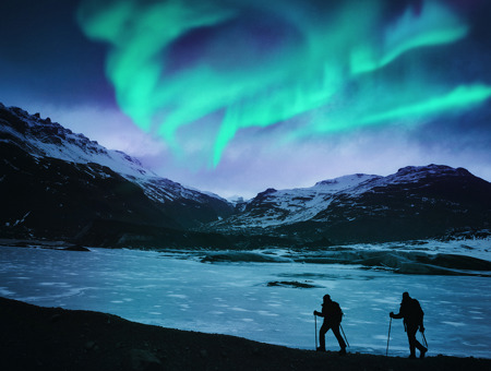 Hikers On A Frozen Lake, Mountains And Northern Lights In The Background