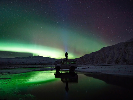 In The Middle Of A Field At Night With Northern Lights And Mountains In The Background